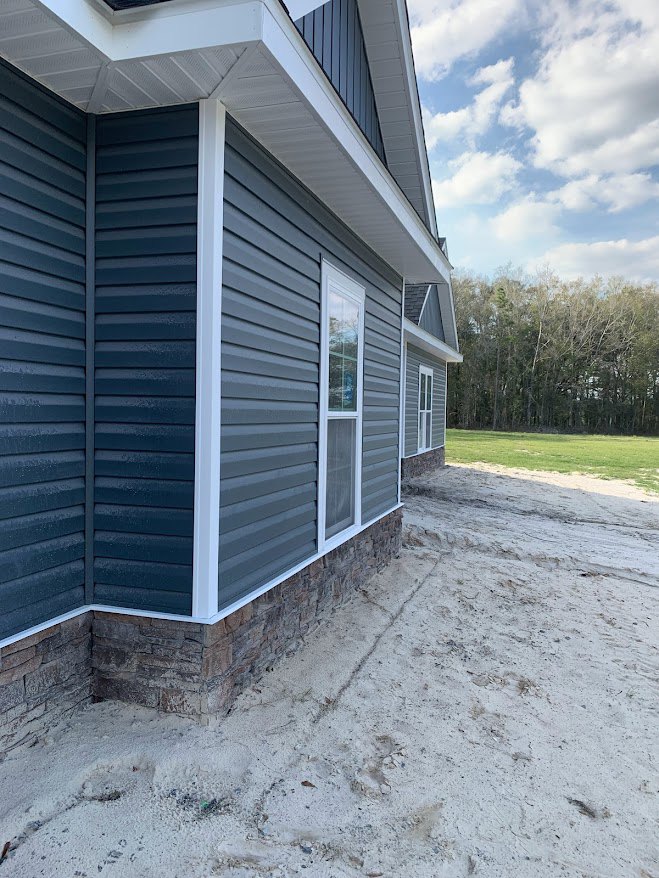 Blue siding house with stone wall, large windows, dirt path leading through grassy yard, mature trees and cloudy sky in background