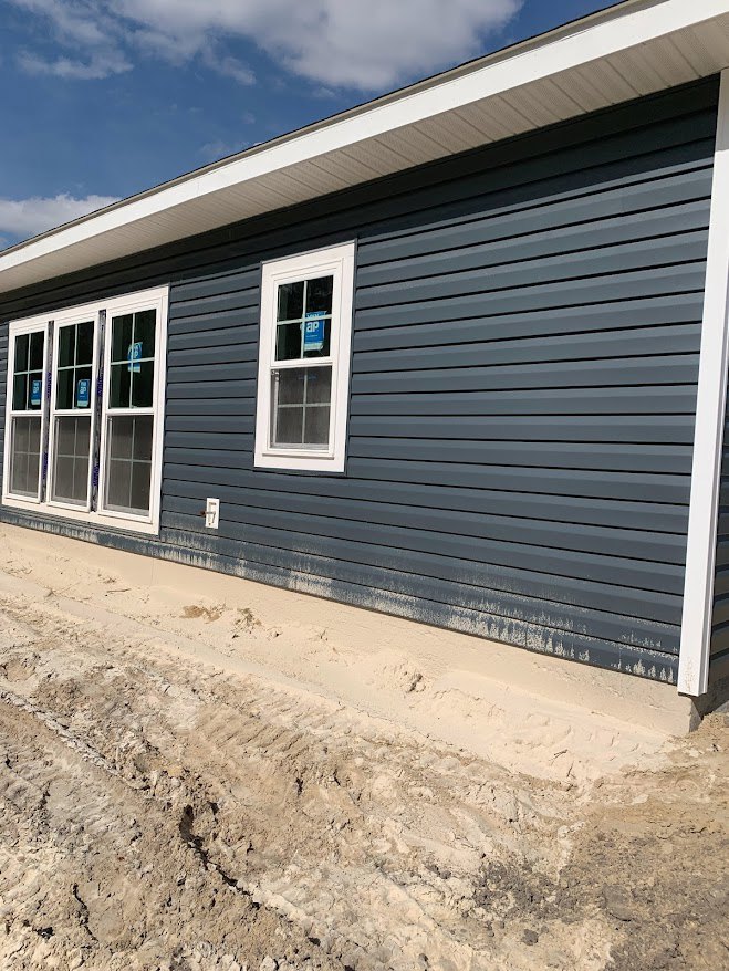 Blue siding and white trim house with multiple windows, sandy ground along the side, dirt road adjacent, cloudy sky overhead