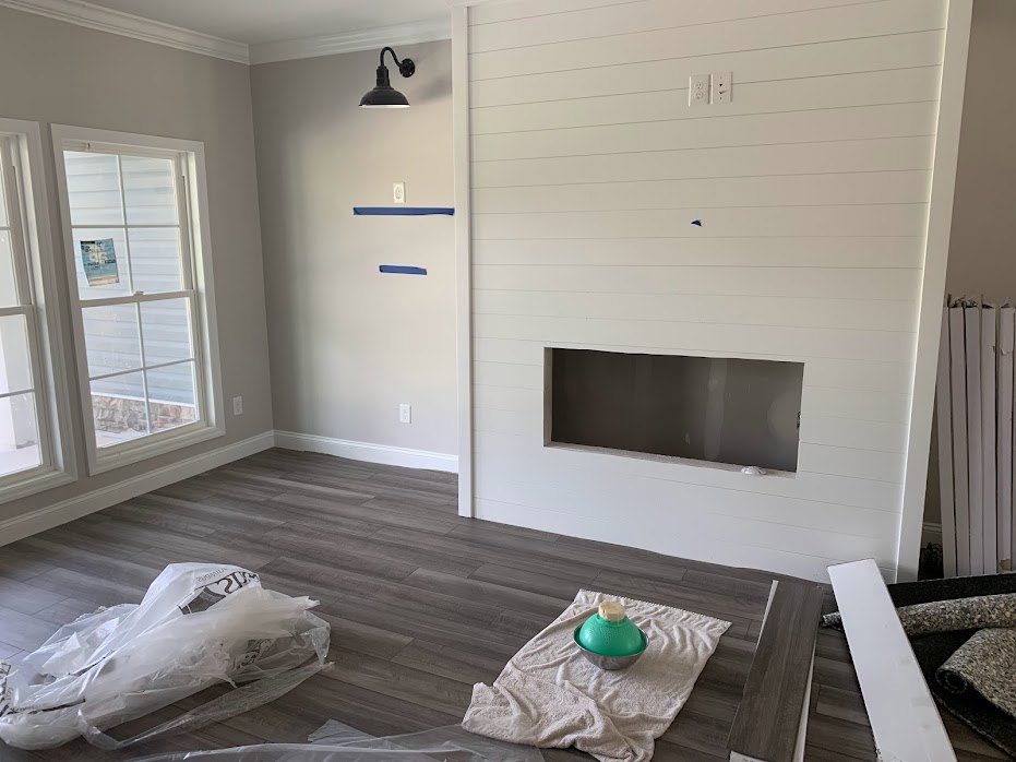 Living room with light wood flooring, white plaster walls, modern black fireplace, curved black light fixture, green bowl with white lid on the floor, plastic bag near wall