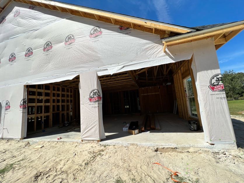Wood-framed house under construction with covered porch, white tarp draped over entry, construction materials on bare ground, trees in background