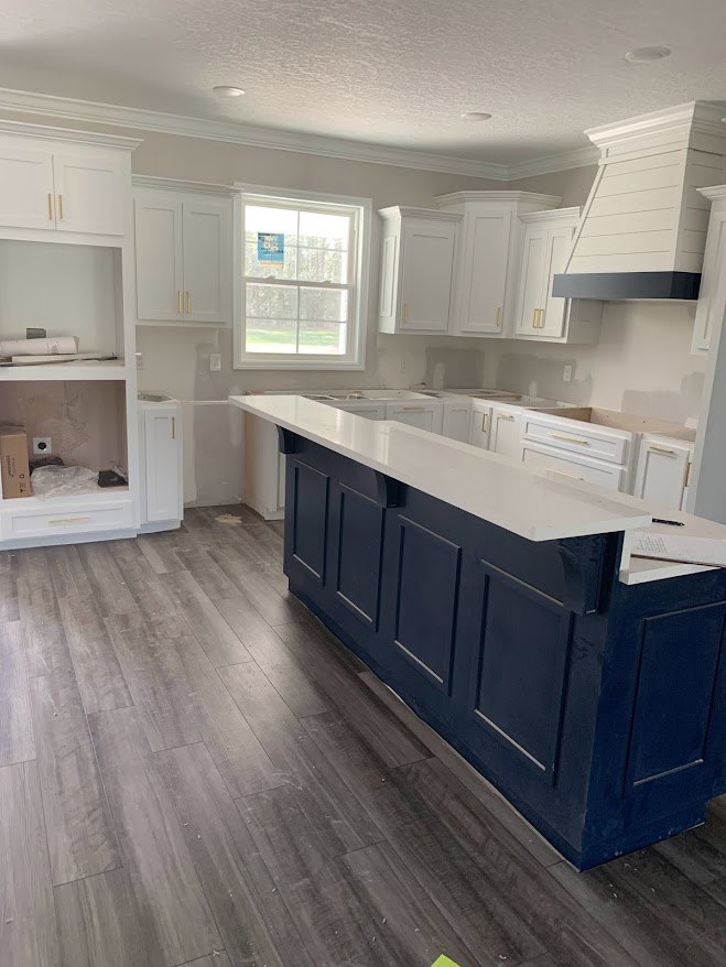 White cabinetry and blue island in kitchen with wood flooring, black and white range hood, stone countertops, window with sign, open shelving holding paper roll