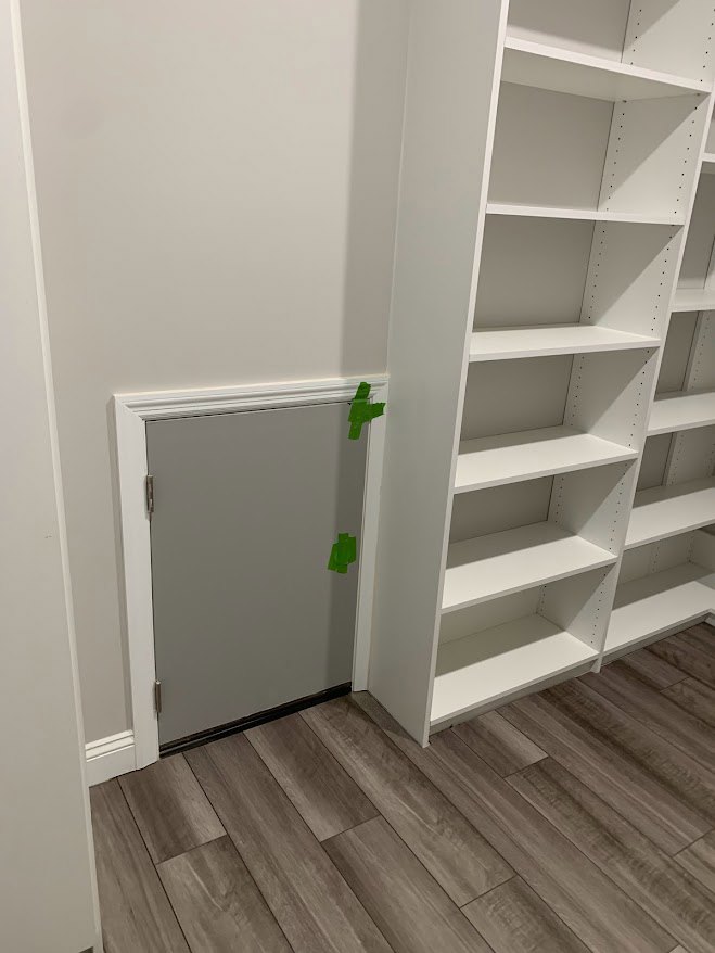 White built-in shelving unit with empty shelves against a plaster wall, adjacent to a grey door marked with green tape, wood flooring and white baseboard visible in foreground.