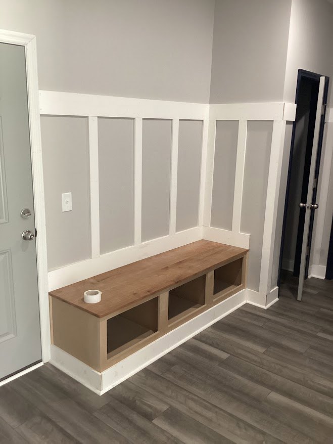 Wooden bench with a roll of tape on top, hardwood plank flooring, white plaster walls, plywood shelf, and a door with a metal handle in a residential room.
