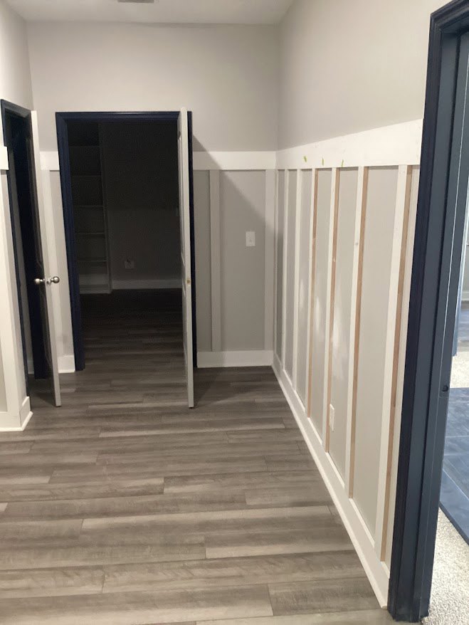 Hallway with light wood flooring, white trim, open door revealing shelving in background, and a light switch on the wall