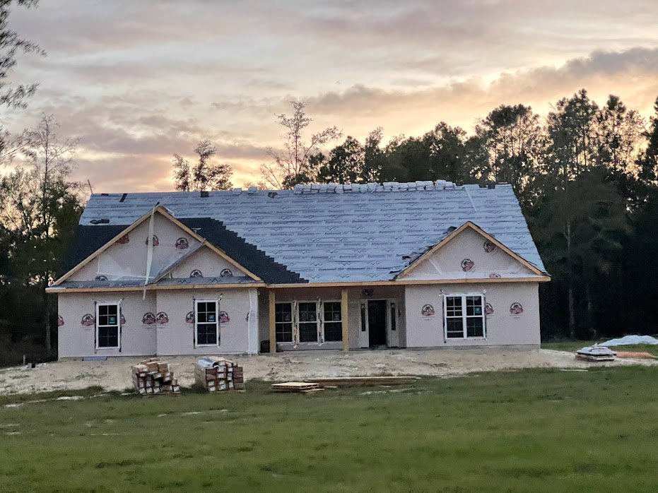 Partially built brick house with multi-pane windows, dark shingle roof, grassy yard scattered with pallets, and mature trees in the background under a cloudy sky