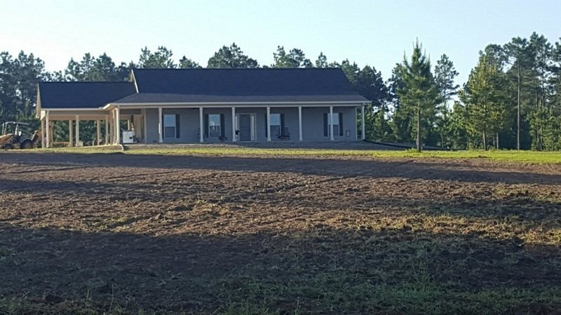 Two-story farmhouse with white siding and covered porch, set beside a large dirt field bordered by grass and trees under a blue sky