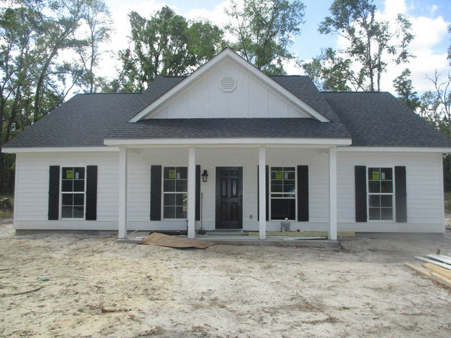 Two-story white house with black shutters and white vent, surrounded by dirt construction area, porch visible, trees in background
