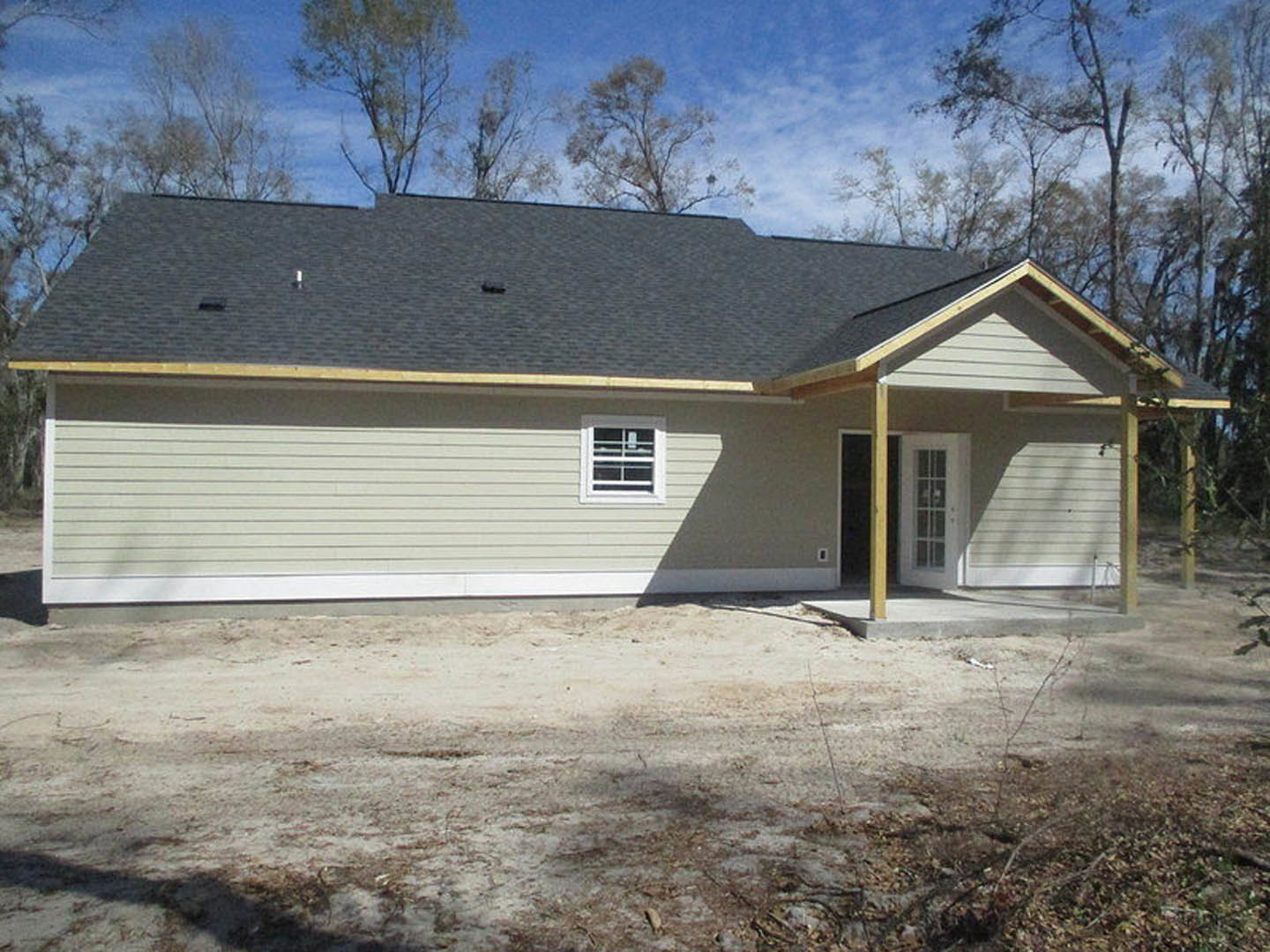 Two-story house with black roof, attached garage, white-framed windows including one with bars, light-colored siding, dirt yard with garden hose, and trees in background