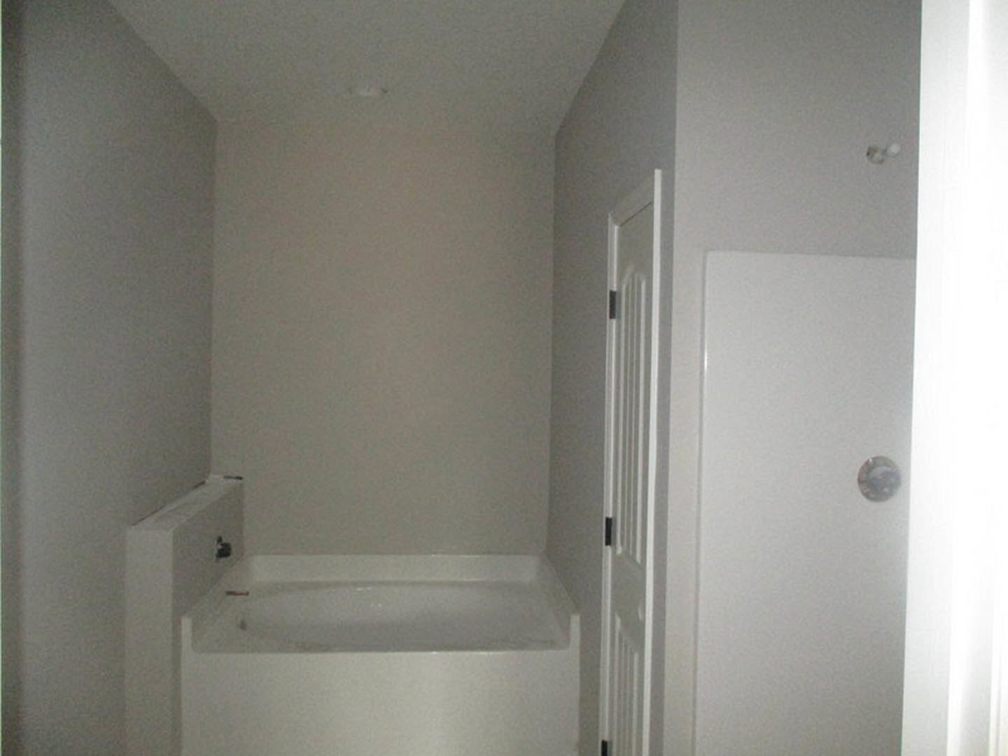 White freestanding bathtub beside a white plaster wall, with a wooden door leading out of the bathroom, chrome plumbing fixtures, and neutral tile flooring