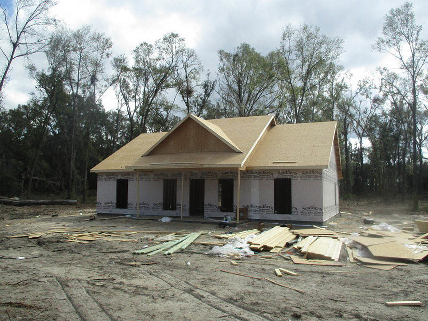 Partially built house with dark framed windows, black exterior walls bordered in white, surrounded by tall trees and piles of lumber on bare ground
