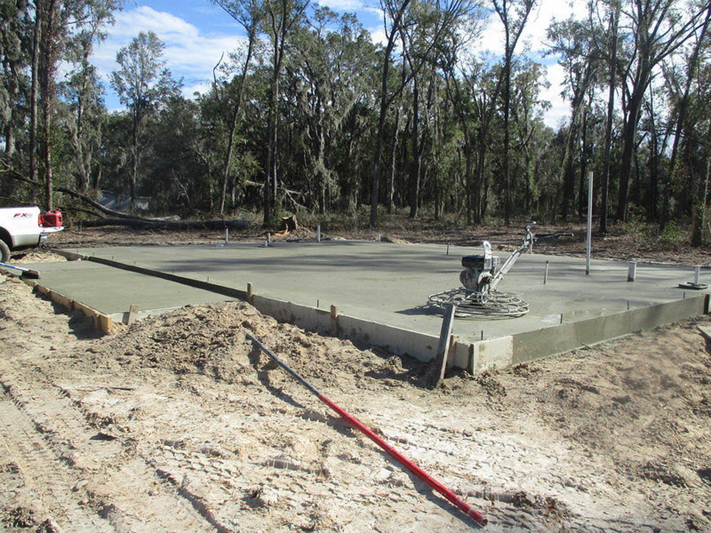 Freshly poured concrete slab surrounded by dense forest, concrete screed machine positioned on the surface, long red and black pole and red pipe lying nearby, white truck with red