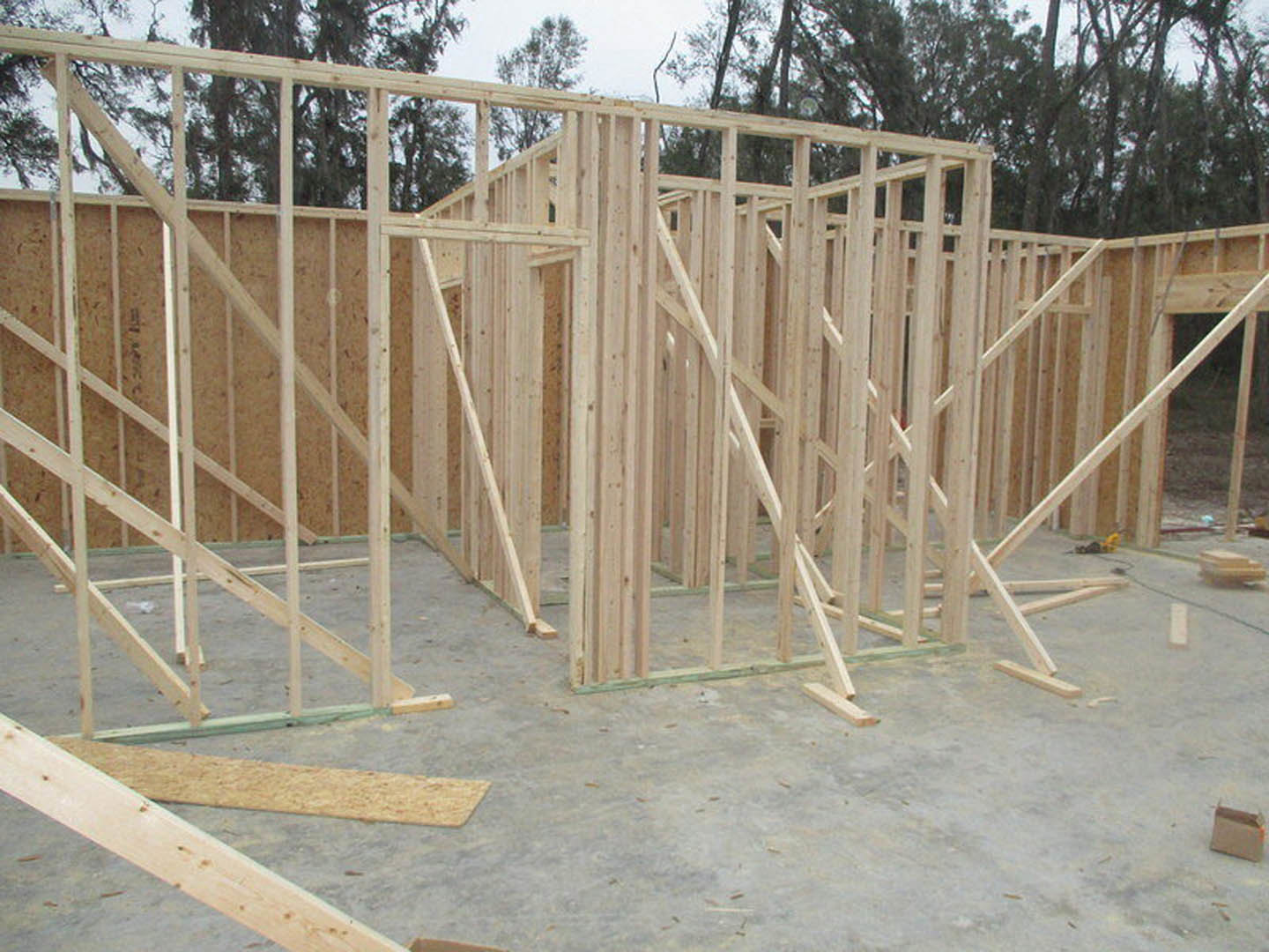 Wooden house frame under construction with exposed lumber beams, surrounded by trees and a car in the background