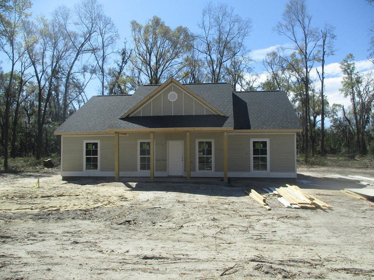 Partially built house with shingled roof, white door with black hardware, wood planks stacked on dirt ground, surrounded by trees, window displaying green sign