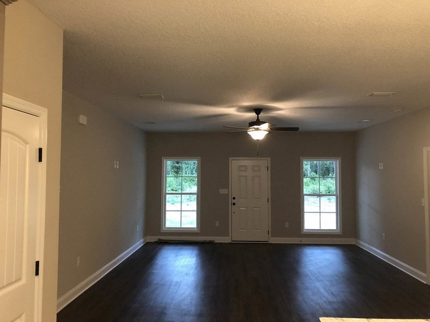 Ceiling fan with light fixture above dark wood laminate flooring, white walls, white door with black knobs, large windows showing leafy trees outside