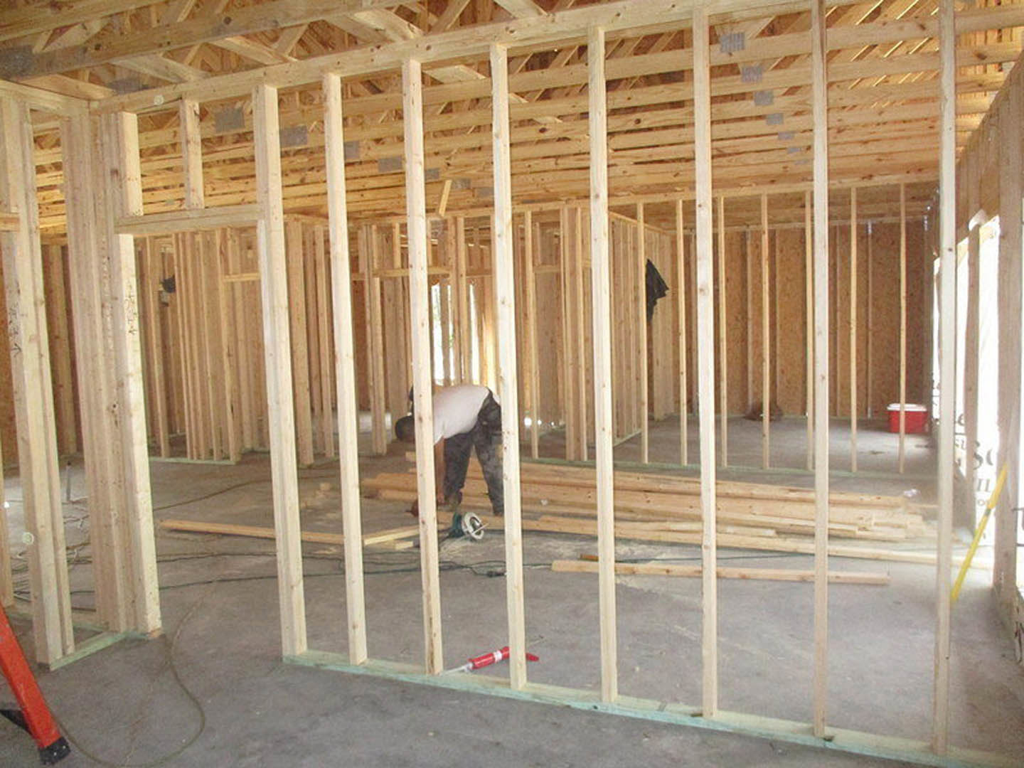Framed wood beams and ceiling insulation in progress, construction worker installing materials on unfinished floor, red container and tools scattered nearby