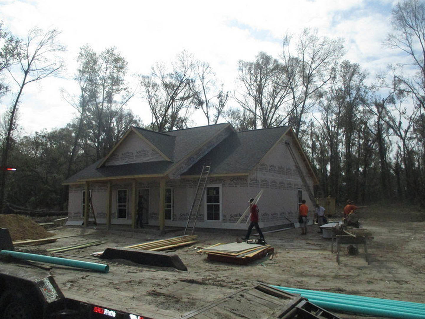 Partially constructed house with exposed framing, workers in red shirts, ladder leaning against exterior, white-framed window, cloudy sky, trees in background
