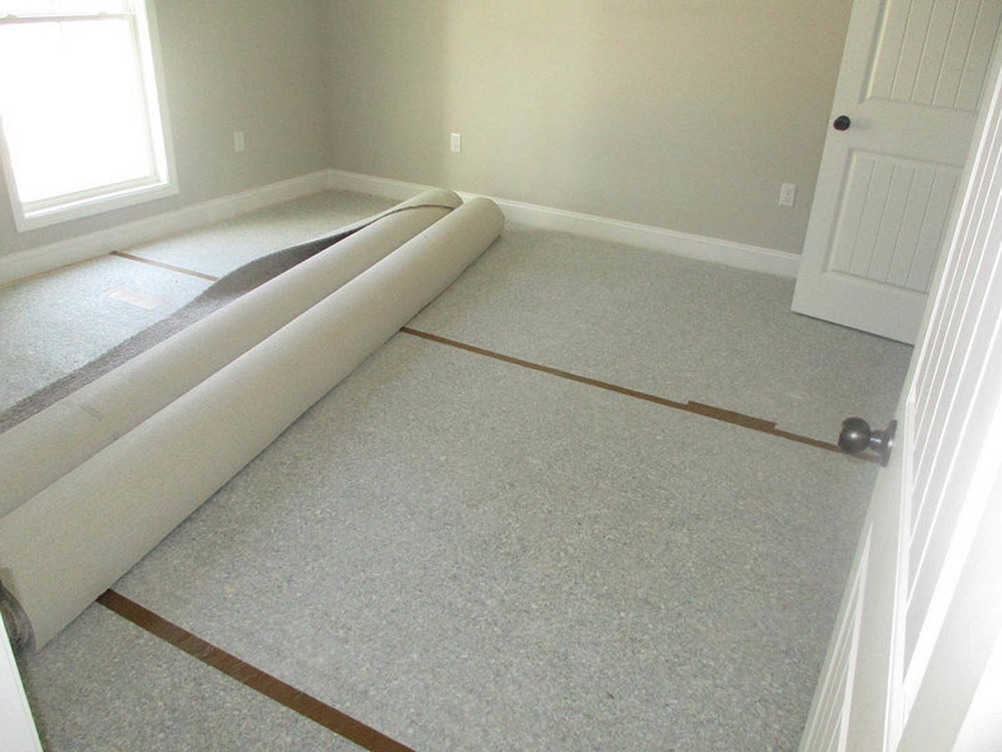 Neutral-toned carpeted floor, white paneled door with matte black handle, large window with white trim, roll of carpet resting near wall, smooth plaster walls, tile flooring