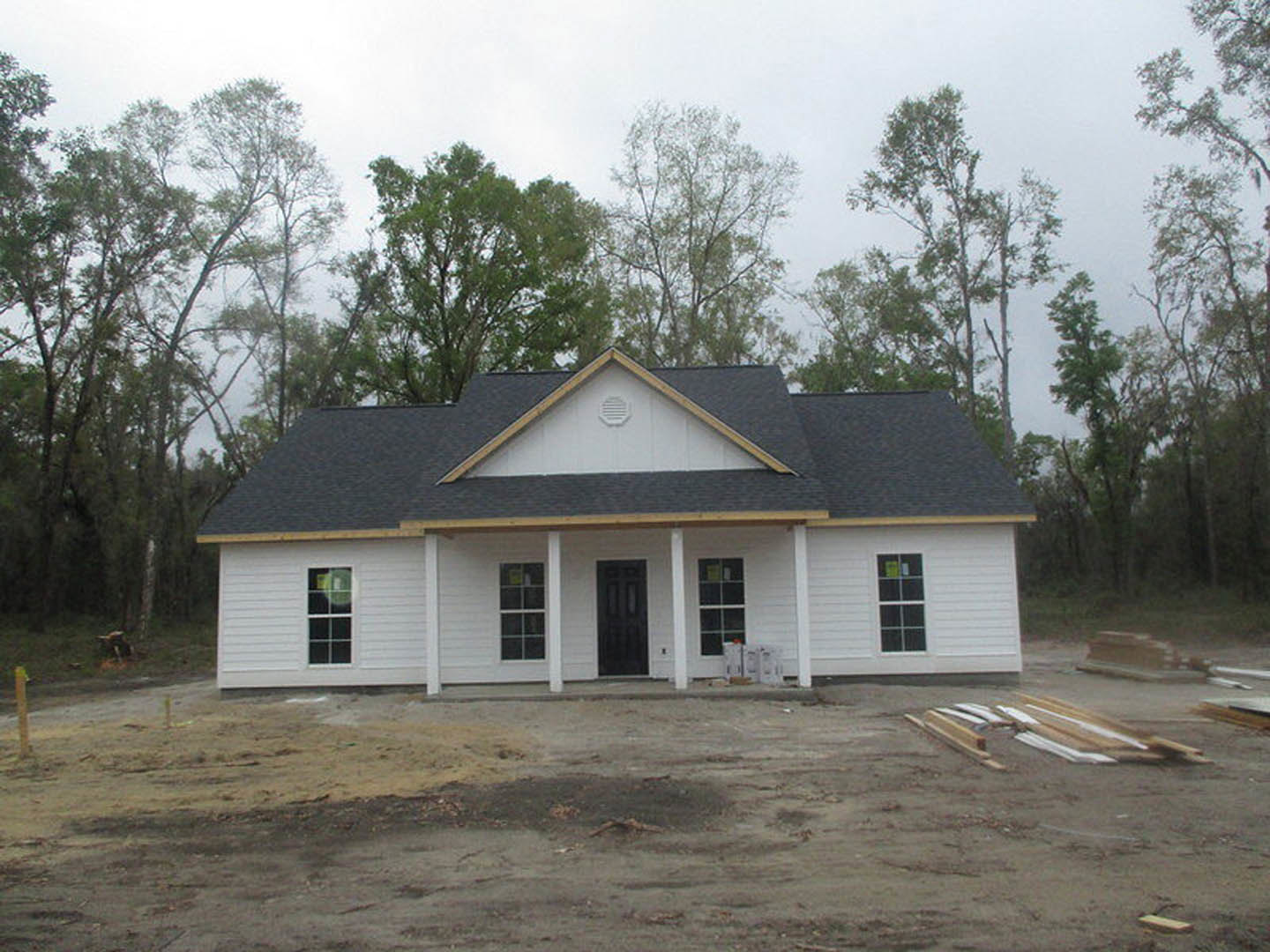White house under construction with black door and white frame, pile of wood on dirt patch in front, Little White House visible in background, trees and sky surrounding property