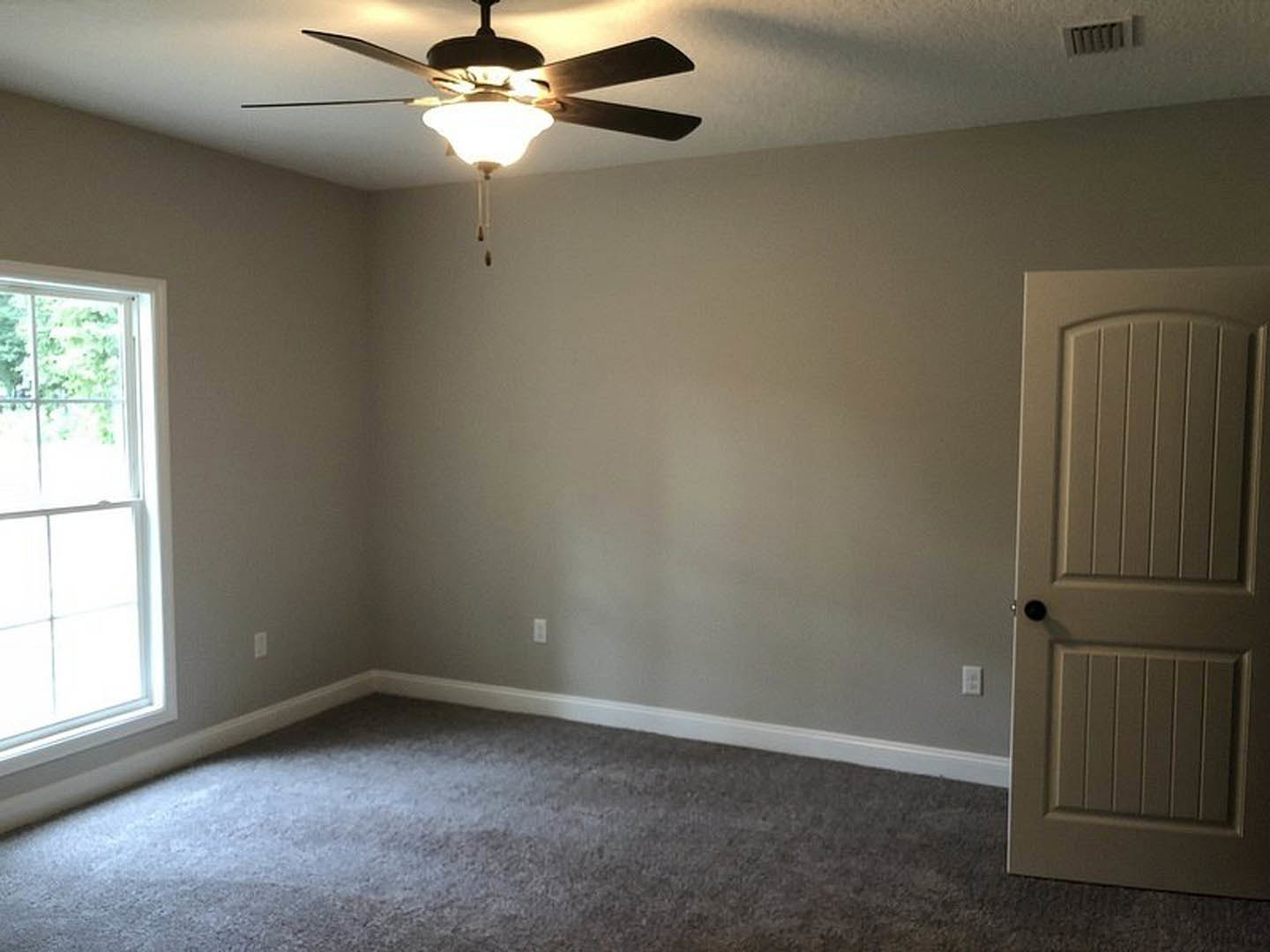 Ceiling fan with light fixture above grey carpeted floor, white door and window on pale walls, wall vent visible