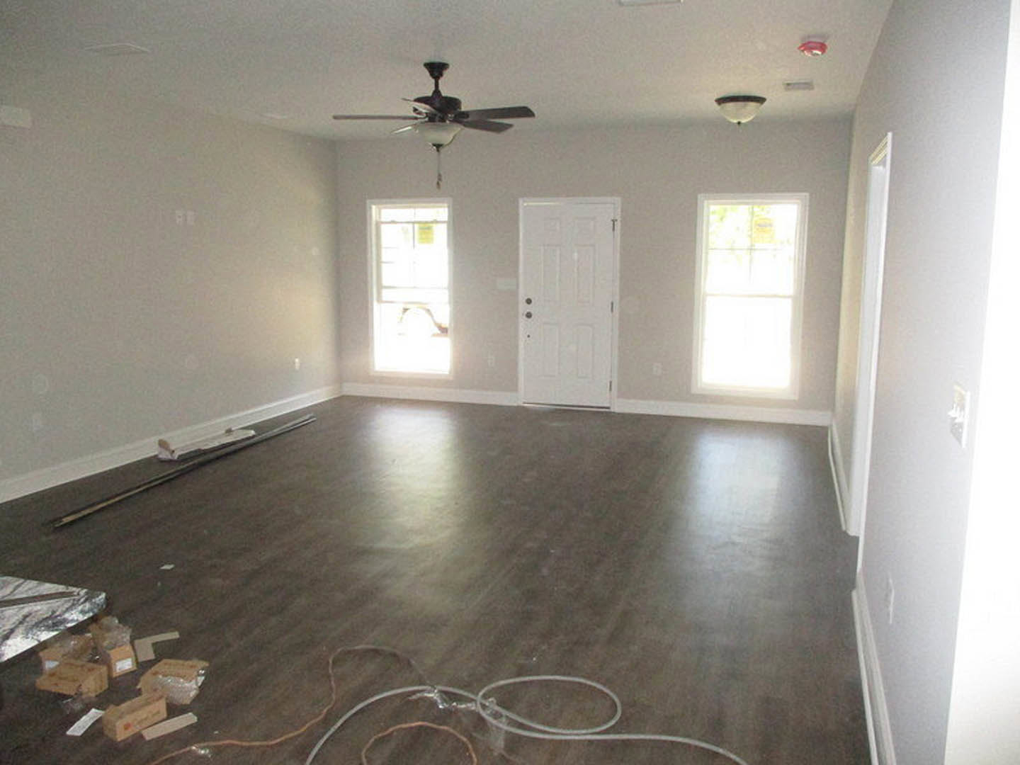 Ceiling fan mounted on white plaster ceiling, wood laminate flooring, white walls, white door with multiple knobs, sunlight streaming through white-framed window