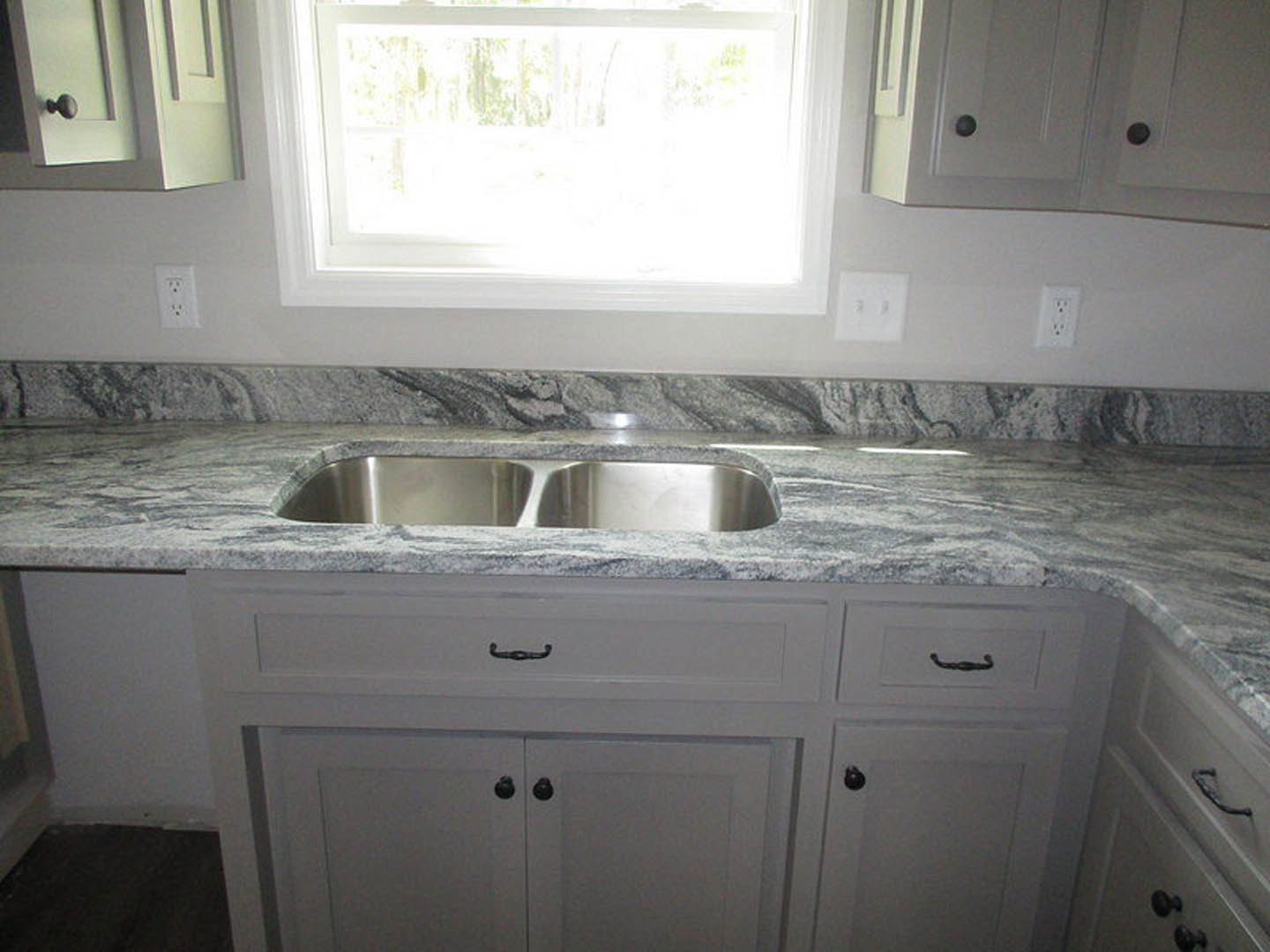 Granite countertop with stainless steel sink, white cabinetry, wall outlet, and sunlit window in a modern kitchen