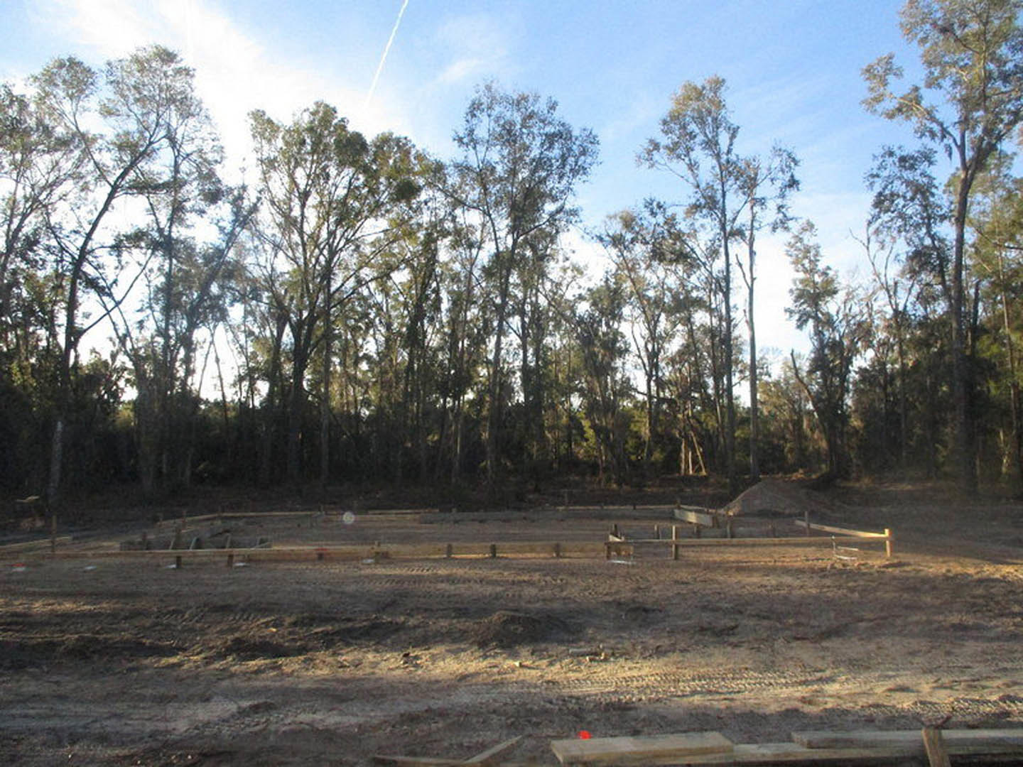 Dirt field bordered by a white fence, group of trees in the background under a partly cloudy sky