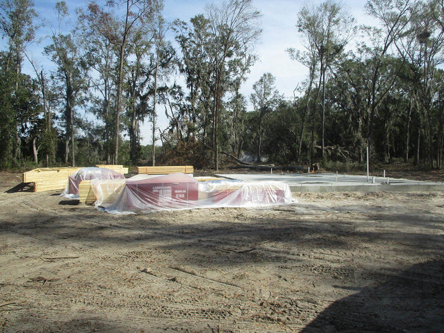 Stacked lumber wrapped in clear plastic beside a red barrel, set outdoors near a covered structure and surrounded by tall leafy trees.
