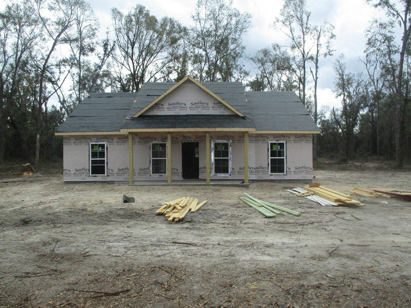Framed house under construction with exposed wood planks, unfinished roof, and a small white cottage visible in the background