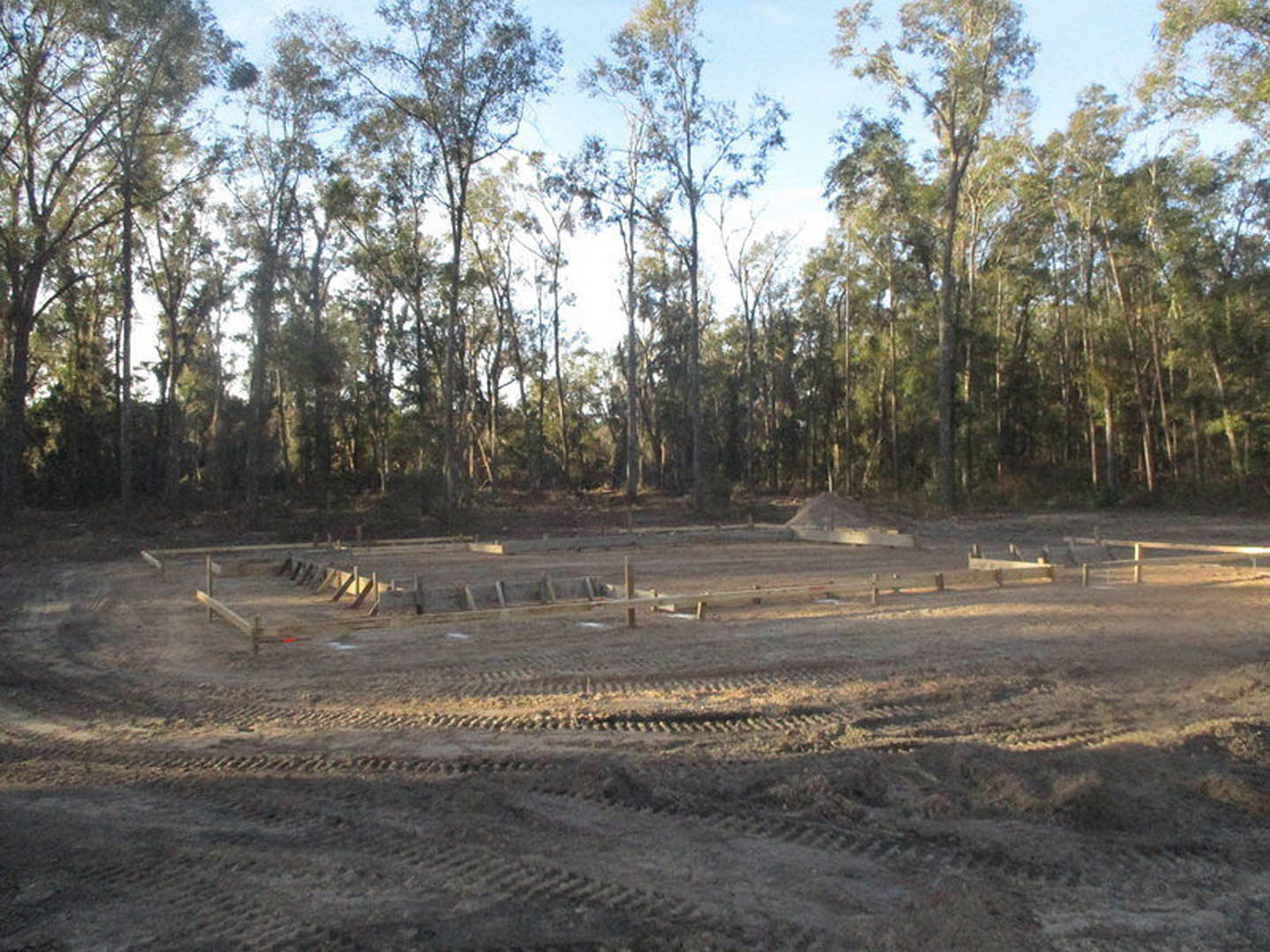 Dirt field with tire tracks, scattered trees in the background, wooden structure partially visible among foliage