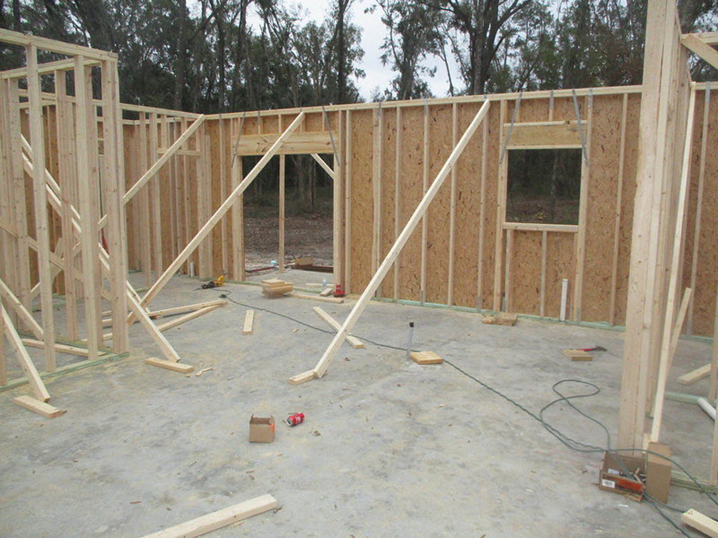 Wood-framed house under construction with exposed beams, lumber planks, and composite materials on the ground; surrounded by trees and construction equipment.