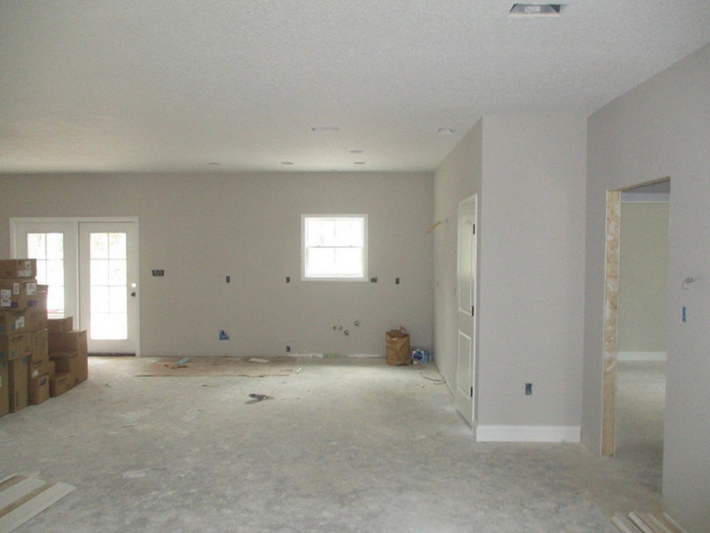 White-walled room with white ceiling, white doors with black handles, brown bag on a white surface, boxes near the wall, close-up of window and wall detailing, light flooring.