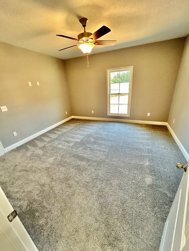 Carpeted room with white plaster walls, ceiling fan with light fixture, open door, and window letting in natural light
