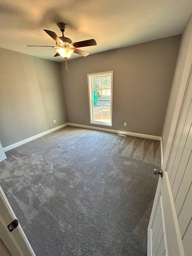 Carpeted room with white plaster walls, ceiling fan with light fixture, large window showing green bin outside