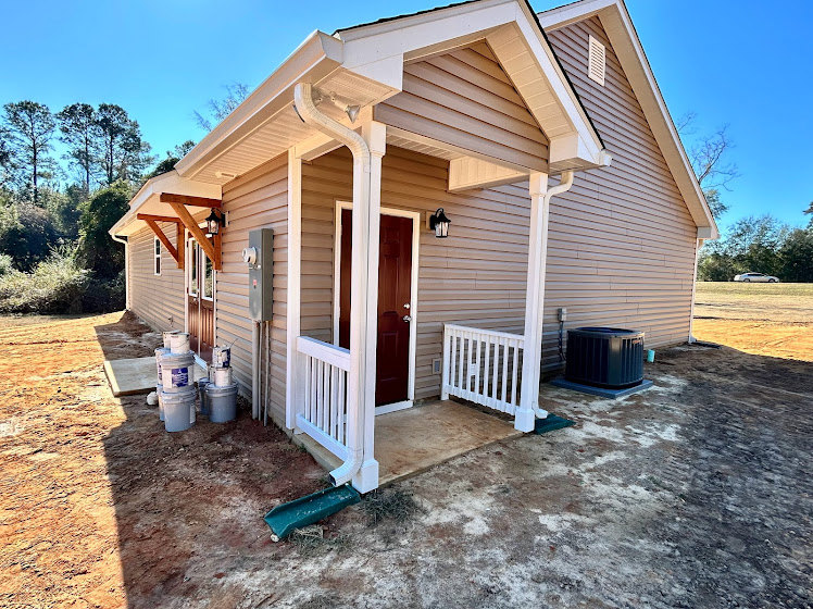 White siding home with covered front porch, wooden door, gabled roof, and landscaped yard under blue sky