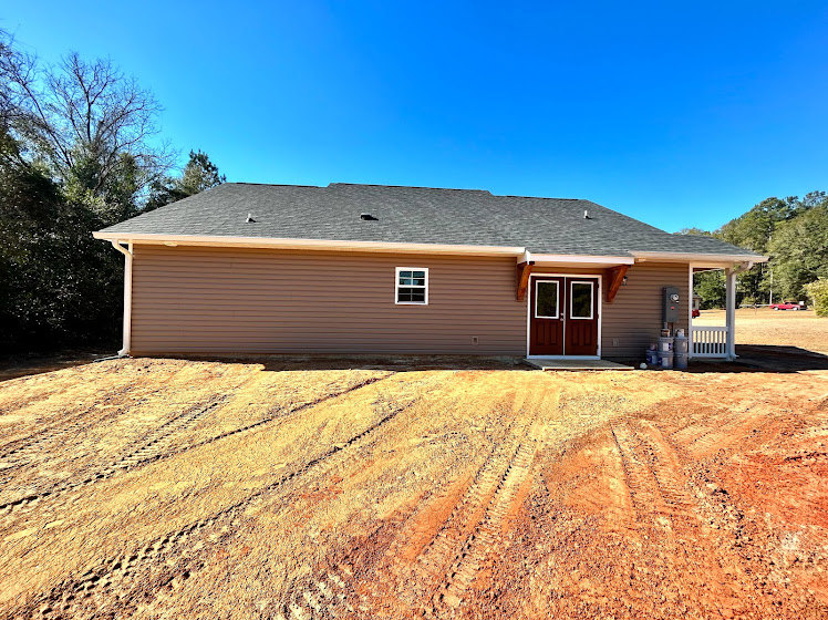 Two-story cottage-style house with light siding, double front doors with glass panels, large windows, and bare dirt yard; surrounded by trees under a clear sky.
