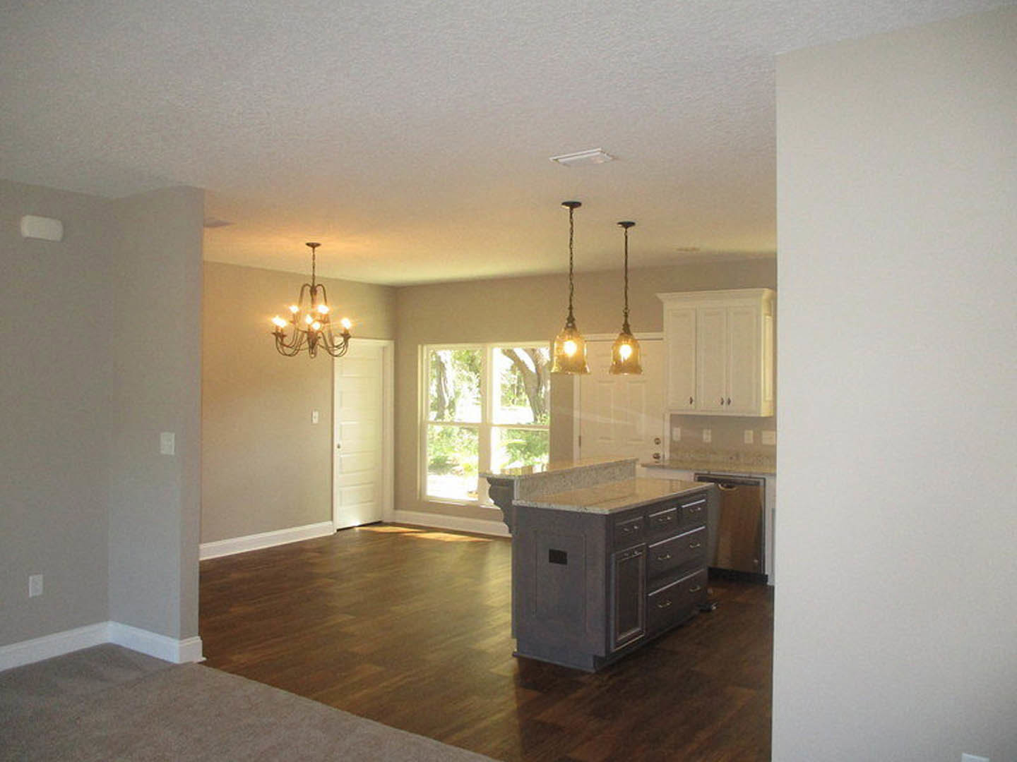Open-concept kitchen and living room featuring wide plank wood flooring, white cabinetry, kitchen island with drawers, modern light fixtures, and a white door with a silver handle