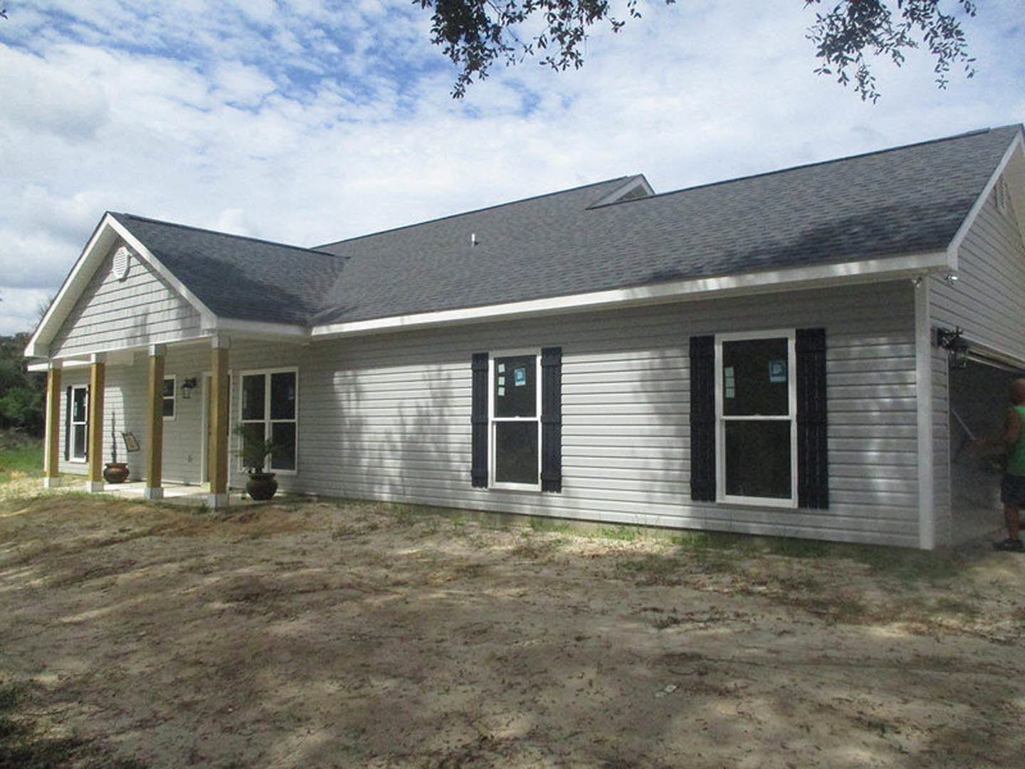 White house with black roof and skylight, white-framed windows, dirt yard in foreground, blue sky with scattered clouds