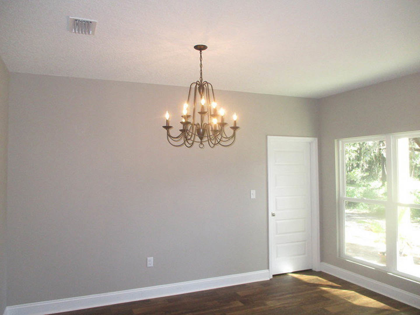 Chandelier with multiple lights hanging from white plaster ceiling, wood floor with white baseboard, white door with silver knob, large window overlooking forest