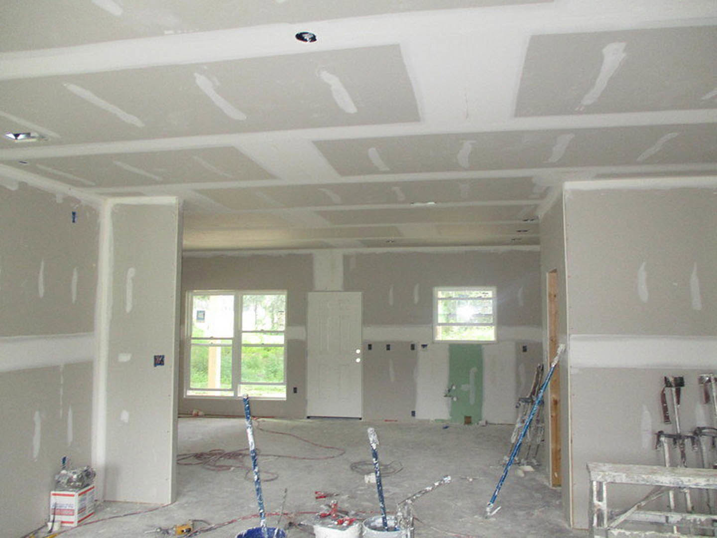 Unfinished room with white plaster walls, paint rollers and cans on the floor, aluminum ladder, window with white frame, white door, blue stick in plastic bag, construction