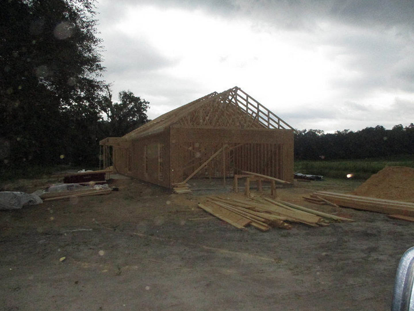 Wood-framed house under construction with exposed beams, unfinished roof, stacked lumber on dirt ground, leafy tree nearby, cloudy sky overhead