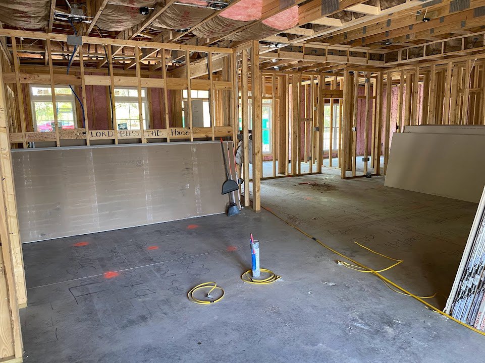 Unfinished room with exposed wooden ceiling beams, insulation, white wallboard with black writing, grey floor, and yellow electrical cable on the ground