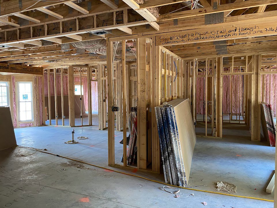 Wood framing and exposed beams inside a residential construction site, stacked lumber on unfinished floor, window with permit sign, partially insulated walls.