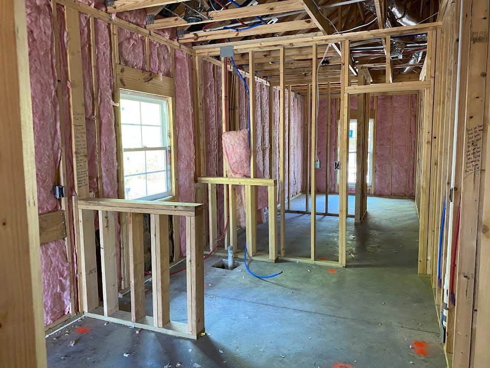 Wood-framed room under construction with exposed beams, white-trimmed window, visible pipes, and blue hose lying on unfinished floor