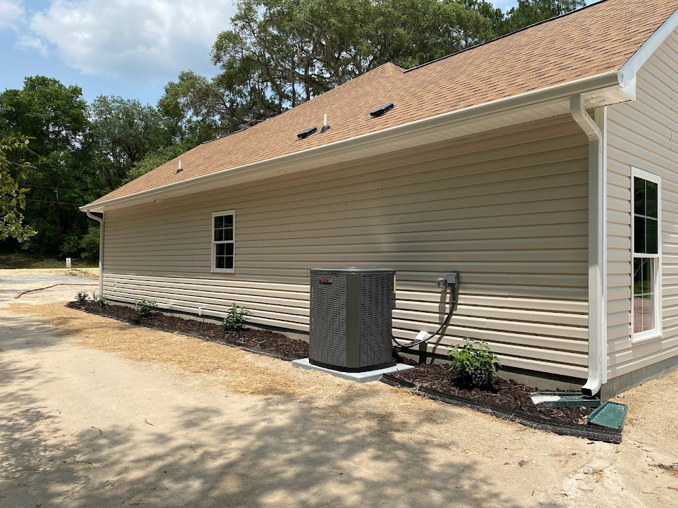 Modern home exterior with grey siding, white-framed windows, large metal heater unit beside driveway, green garden plants, and cloudy sky with trees in background