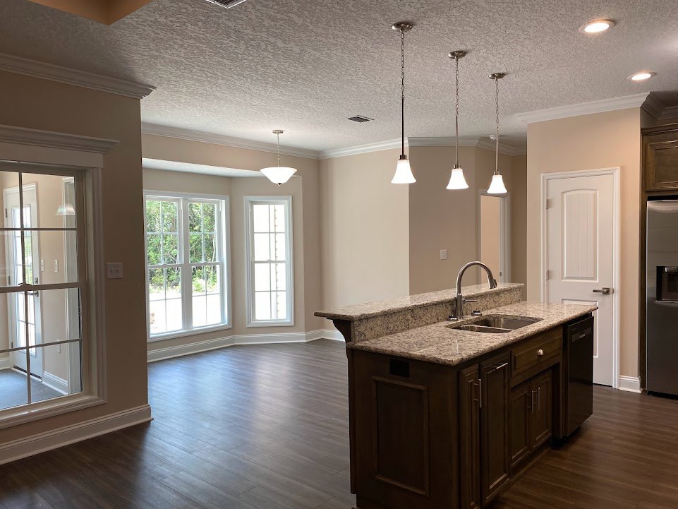 Open kitchen with wood flooring, central island featuring a built-in sink, white cabinetry, stone countertops, pendant lighting, large window with white frame overlooking trees
