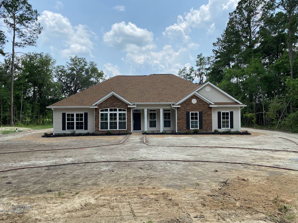 Two-story home with brown roof, large dirt driveway, prominent window, surrounded by tall trees under a partly cloudy blue sky