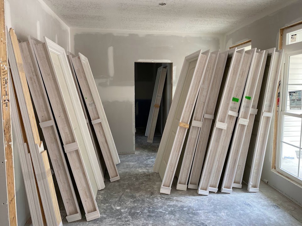 Unfinished room with stacked white doors, white door frame, white wood planks, window displaying sign, and white ladder against plastered wall