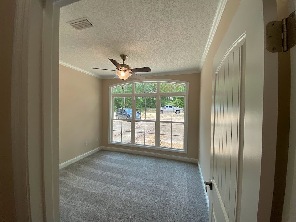 Carpeted room with white vertical blinds, multiple windows, ceiling fan with light, metal hinge on wall, and parked car visible outside.