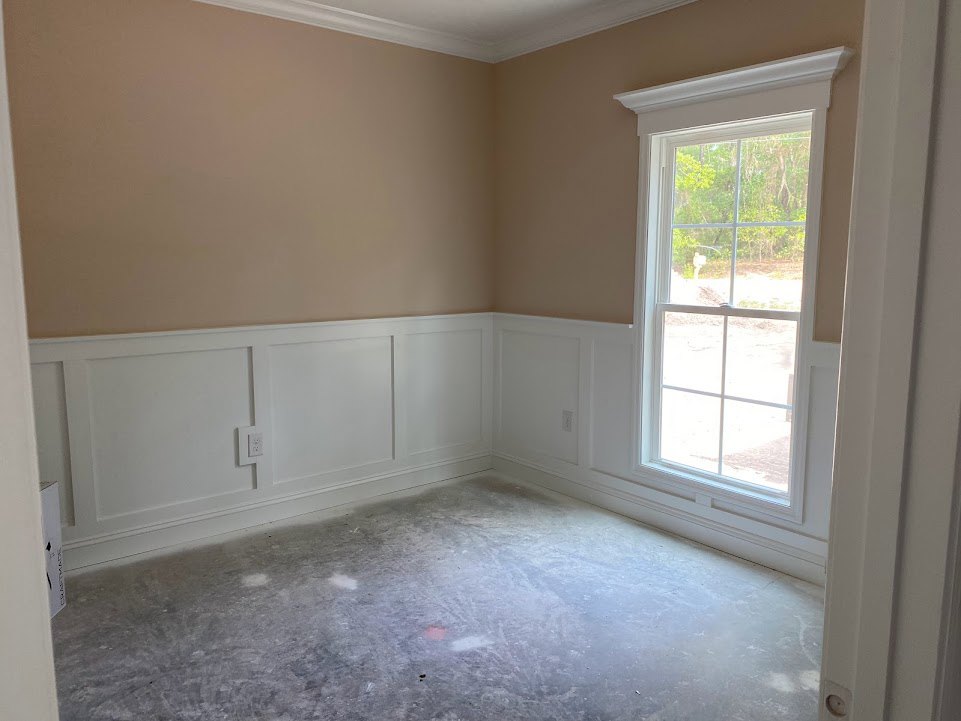 Bright room featuring a large window overlooking trees, concrete floor painted white, white paneled walls, and a white ceiling with brown accent surface
