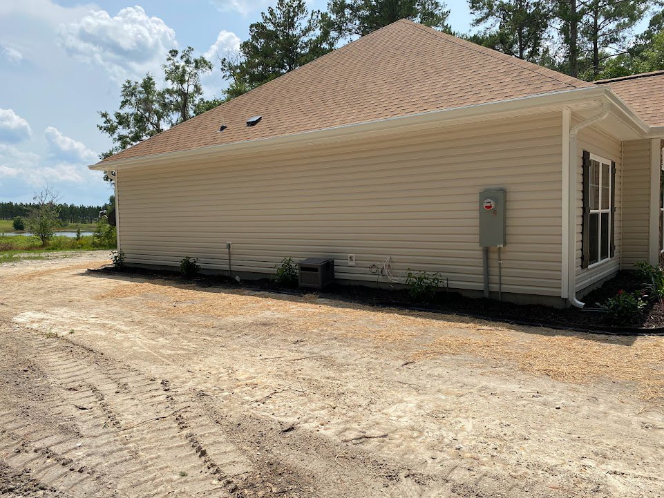 Modern home with light gray siding, large windows, and a black utility box mounted on the exterior wall; dirt road runs alongside the house, surrounded by trees and plants under a